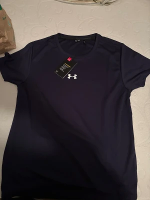 Under armour tröja - Snygg mörkblå t-shirt från Under Armour Project Rock-kollektionen. Tillverkad i ett lätt och snabbtorkande funktionsmaterial, perfekt för träning. Klassisk passform med rund hals och korta ärmar. Ikonisk Project Rock-logga på ryggen och UA-logga på bröstet.