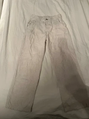 Beiga linnebyxor med raka ben - Säljer ett par ljusbeiga linnebyxor med raka ben och hög midja. Byxorna har knapp och dragkedja framtill, samt resår baktill i midjan för extra komfort. Perfekta för en avslappnad och stilren look.