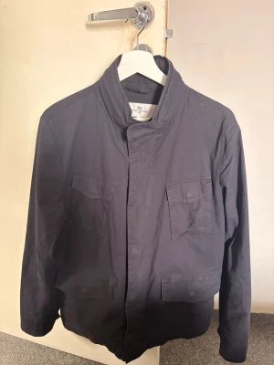 Mörkblå jacka från John Henric - Snygg mörkblå jacka från John Henric med hög krage, dragkedja och tryckknappar. Två bröstfickor med lock och knapp, samt diskreta sidofickor. Jackan har en stilren och minimalistisk design, perfekt för blåsiga dagar. Materialet känns lätt och smidigt.