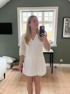 Vit kort klänning med broderad spets - Söt vit kortklänning med v-ringning och halvlånga ärmar. Klänningen har ett broderat spetsmönster med blommiga detaljer och är tillverkad i bomull. Perfekt för sommarens alla tillfällen!