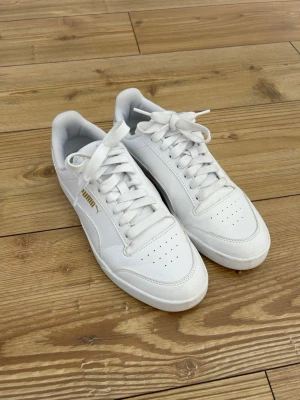 Vita Puma sneakers - Fräscha vita sneakers från Puma med klassisk låg profil och guldfärgad logga på sidan. Perfekta för en clean och stilren look.