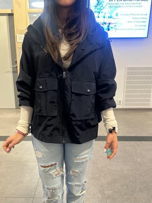 Svart vindjacka med huva - Snygg svart vindjacka med dragkedja och stor huva. Jackan har två stora fickor med lock och knapp framtill. Rak passform och något kortare modell som ger en trendig look. Perfekt för blåsiga dagar och enkel att matcha med olika outfits.