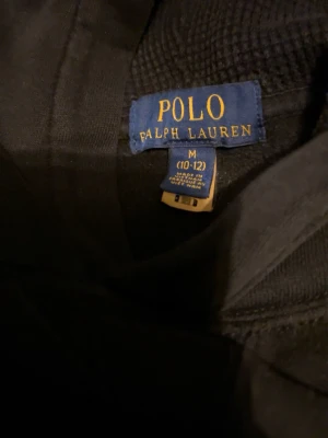 Svart tröja från Polo Ralph Lauren - Svart hoodie från Polo Ralph Lauren med den klassiska röda broderade loggan på bröstet. Tröjan har en avslappnad passform och är tillverkad i mjukt material som känns skönt mot huden. Perfekt för dig som gillar stilrena och tidlösa plagg.