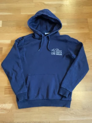 Mörkblå hoodie från Les Deux - Mörkblå hoodie från Les Deux med vit logga tryckt på bröstet. Klassisk känguruficka framtill, justerbar dragsko i huvan och ribbade muddar vid ärmslut och nederkant. Perfekt för en avslappnad och stilren look.