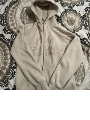 Beige zip-hoodie från Peak Performance - Säljer en beige hoodie från Peak Performance med dragkedja och huva. Tryck på bröstet med texten 'Peak Performance Since 1986'. Dragkedjan är av men annars är den perfekt. Perfekt för en chill och avslappnad stil.