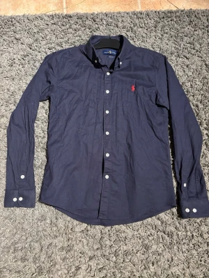 Marinblå skjorta från Ralph Lauren - Klassisk marinblå skjorta från Ralph Lauren i modellen Classic Fit. Skjortan har långa ärmar, button-down krage och vita knappar .                               OBS! Det är en M men sitter som S. Hör av er om ni har funderingar👍