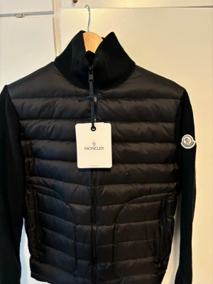 Svart dunväst från Moncler äkta - Snygg svart dunväst från Moncler med stickade svarta ärmar och klassisk Moncler-logga på armen. Västen har två dragkedjeförsedda fickor framtill och quiltad design. Utan luva. Och priset diskuteras 