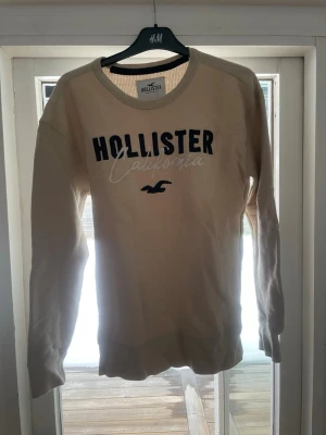 Beige stickad tröja Hollister - Stickad beige tröja från Hollister med rund halsringning och svart broderad text 'HOLLISTER California' på bröstet. Tröjan har långa ärmar och ribbade muddar vid ärmslut och nederkant. Perfekt för en avslappnad och stilren look. Jag är 186 väger 69kg sitter kanon på mig!