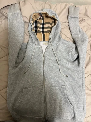 Grå zip kofta Burberry - Säljer en grå zip hoodie från Burberry med klassiskt beige rutigt foder i huvan. Hoodien har dragsko i huvan, två stora fickor fram och ribbade muddar vid ärmar och nederkant. Perfekt för dig som gillar stilrena och exklusiva plagg.