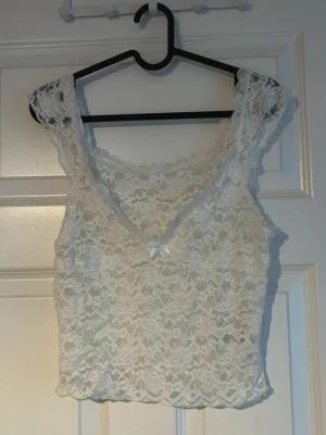 Vit spetstopp med rosett - vit spetstopp med v-ringning och söt liten rosett framtill 🤍 Transparent spetsmönster som ger ett feminint och elegant uttryck. Supersnygg till höga jeans eller en kjol för en mjuk, trendig look.