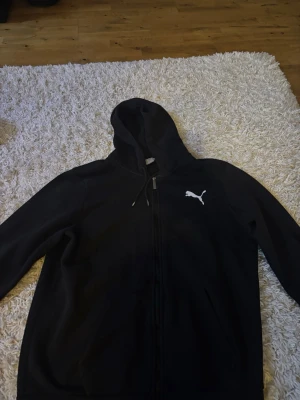 Svart hoodie med dragkedja från Puma - Säljer en svart hoodie från Puma med dragkedja framtill och vit logga på bröstet. Hoodien har justerbar huva med snören och två fickor fram. Perfekt för en avslappnad och sportig stil.