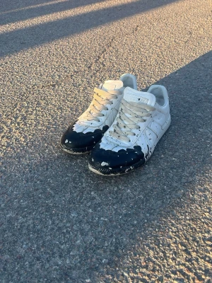 Vita Gats med svart drip - Gats,  Maison Margiela med svart drip-effekt över tån och sidorna. Skorna har klassisk låg siluett, vita skosnören och är tillverkade i läder. Perfekt för dig som vill sticka ut med en kreativ och edgy look. Skriv för funderingar pris ej hugget i sten👍