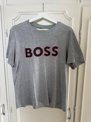 Grå BOSS t-shirt med tryck - Snygg grå t-shirt från BOSS med stort svart och rött BOSS-tryck på bröstet. Klassisk rund halsringning och korta ärmar. Tillverkad i mjuk bomull som känns skön mot huden. Perfekt för en avslappnad och stilren look.