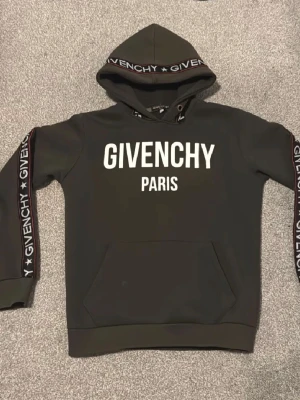 Givenchy hoodie - Svart hoodie från Givenchy med stor vit logga och texten 'PARIS' på bröstet. Huva med snörning och Givenchy-tryck längs ärmarna och huvkanten. Känguruficka framtill och ribbade muddar. Perfekt för en trendig streetwear-look.