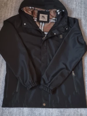 Svart vindjacka från Burberry - Stilren svart vindjacka från Burberry med klassiskt rutigt foder i beige, svart, vitt och rött. Jackan har huva, dragkedja, tryckknappar och två fickor med dragkedja. Elastiska muddar vid ärmslut och diskret Burberry-logga på bröstet. XXL men sitter mer som en XL