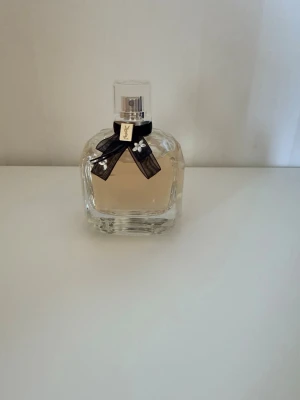 Yves Saint Laurent Mon Paris edp 90ml - Elegant parfym från Yves Saint Laurent, Mon Paris, i en genomskinlig glasflaska med ljusrosa doft. Flaskan har en dekorativ svart rosett med små pärlor och metallbricka vid halsen. Klassisk och lyxig design som passar perfekt på sminkbordet.