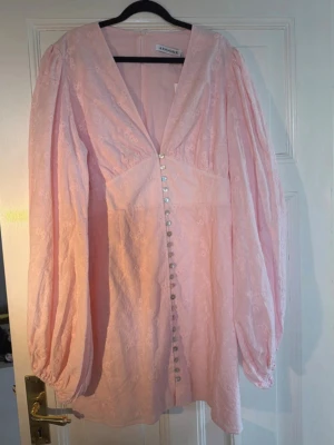 Ljusrosa Florence klänning från Adoore  - Supersöt ljusrosa kortklänning från Andohé med v-ringning, långa puffärmar och dekorativa knappar framtill. Klänningen har ett subtilt blommigt mönster och är gjord i ett tunt, lätt material som känns luftigt och romantiskt.