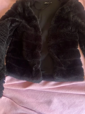 Svart fuskpälsjacka från Gina Tricot - Mysig svart fuskpälsjacka från Gina Tricot med mjukt foder och öppen front. Jackan har en klassisk rak passform och långa ärmar, perfekt för att hålla värmen under kalla dagar. Snygg och trendig look som passar till många outfits.