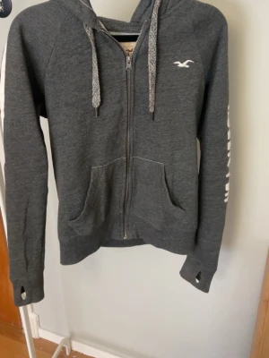 Mörkgrå zip hoodie från Hollister - Mörkgrå hoodie med dragkedja från Hollister. Har vita detaljer som logga på bröstet och text på ärmen, samt två fickor fram och huva med dragsnören. Tillverkad i mjukt sweatshirtmaterial och har tumhål vid ärmsluten. Perfekt för en avslappnad stil.
