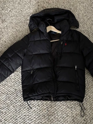 Svart pufferjacka från Polo Ralph Lauren - Snygg svart pufferjacka från Polo Ralph Lauren med huva och dragkedja framtill. Jackan har två framfickor med dragkedja och den klassiska röda loggan broderad på bröstet. Perfekt för kalla vinterdagar och riktigt skön att bära.