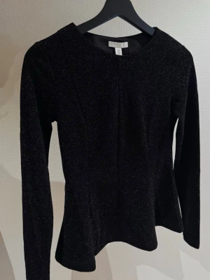 Svart glittrig topp från H&M - Säljer en svart långärmad topp från H&M med glittrigt tyg och figurnära passform. Toppen har rund halsringning och lätt utsvängd nederdel som ger en snygg siluett. extra.