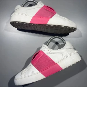 Valentino vita sneakers med rosa band - Snygga vita sneakers från Valentino Garavani med breda rosa band över vristen. Skorna har en chunky sula och detaljerade nitar baktill. Tillverkade i läder med cool design som sticker ut. Perfekta för dig som vill ha något unikt och trendigt.