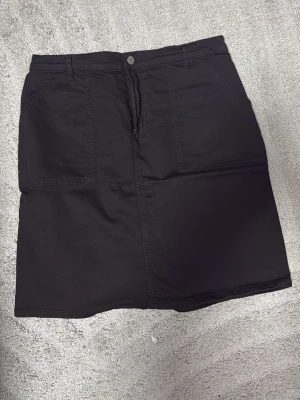 Svart jeanskjol med fickor - Säljer en svart jeanskjol med raka linjer och två stora fickor framtill. Kjolen har knapp och dragkedja i midjan samt hällor för skärp. Materialet är kraftigt och ger en klassisk denimkänsla. Perfekt basplagg till garderoben.