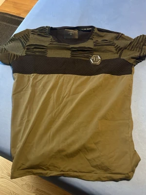 Grön Philipp Plein t-shirt med detaljer - Cool grön t-shirt från Philipp Plein med svarta ribbade och slitna detaljer upptill samt en metallisk logga på bröstet. T-shirten har rund hals och korta ärmar. Perfekt för dig som vill sticka ut med en unik design.