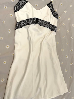 Vitt nattlinne med spetsdetaljer - Snyggt vitt nattlinne med tunna axelband och svarta spetsdetaljer vid byst och midja. Materialet är satin och den är mjukt och bekväm. Perfekt för dig som vill ha något bekvämt men ändå stilrent till natten.