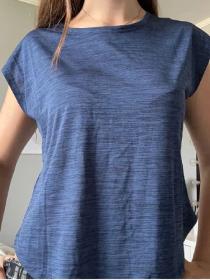 Blå loose-fit sporttopp - Säljer en blå loose-fit sporttopp med korta ärmar och rund hals. Materialet är lätt och andas, perfekt för träning eller aktiva dagar. T-shirten har en diskret melerad effekt och en avslappnad passform som ger bra rörelsefrihet.