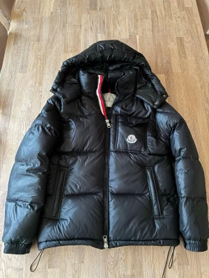 Moncler jacka - Snygg svart dunjacka från Moncler med glansig finish, stor huva och dragkedja med rödvit detalj. Jackan har två framfickor med dragkedja och en bröstficka med Moncler-logga. Perfekt för kalla dagar och riktigt trendig stil.