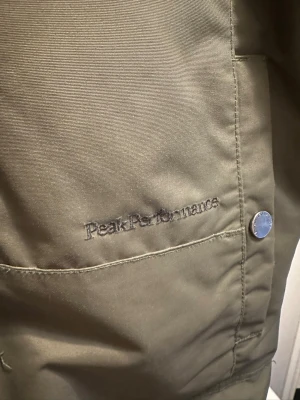 Olivgrön parkas Peak Performance - Snygg olivgrön parkas från Peak Performance med huva, dragkedja och tryckknappar framtill. Jackan har flera praktiska fickor och diskret broderad logga på bröstet. Perfekt för kalla dagar och riktigt stilren look.