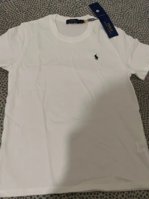 Vit t-shirt från Polo Ralph Lauren - Klassisk vit t-shirt från Polo Ralph Lauren med rund halsringning och den ikoniska lilla logotypen broderad på bröstet. Tillverkad i mjuk bomull för en skön och stilren look. Perfekt basplagg till garderoben.