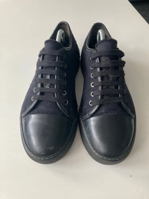 Svarta sneakers med mockadetaljer - Stilrena svarta sneakers med ovandel i mocka och tåparti samt sula i slätt svart läder. Klassisk låg modell med svarta skosnören och metallöljetter. Perfekta för en clean och enkel look.