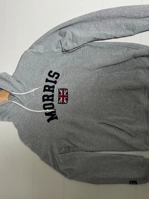 Grå hoodie från Morris  - Snygg grå hoodie från Morris med svart broderad text och en Union Jack-flagga på bröstet. Klassisk känguruficka framtill och vita dragsnören i huvan. Tillverkad i mjuk bomullsblandning, perfekt för en avslappnad stil. Storlek M