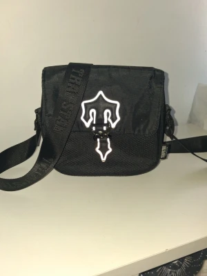 Svart axelväska från Thrasher - Snygg svart axelväska från Thrasher med justerbar rem och ett reflekterande logomärke på framsidan. Väskan har en fyrkantig form och är tillverkad i slitstarkt tygmaterial. Perfekt för att bära det viktigaste och ger en streetwear vibe.