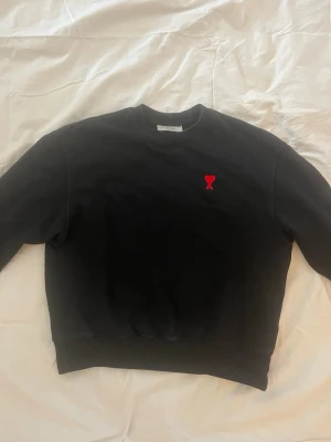 Svart sweatshirt från Ami Paris - Svart sweatshirt från Ami Paris med röd broderad logga i form av ett hjärta på bröstet. Klassisk rund halsringning och ribbade muddar vid ärmslut och nederkant. Perfekt för en clean och stilren look.