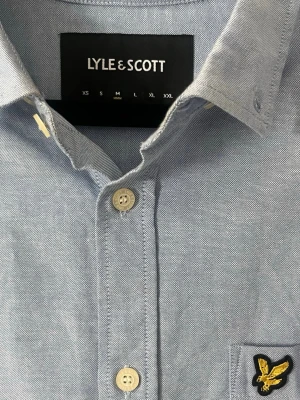 Ljusblå skjorta från Lyle & Scott - Klassisk ljusblå skjorta från Lyle & Scott med button-down krage och bröstficka med den ikoniska gula loggan. Skjortan har långa ärmar och vita knappar framtill. Perfekt för en clean och stilren look.