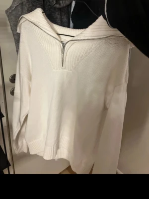 Vit stickad tröja med dragkedja - Vit stickad tröja med bred krage och dragkedja framtill. Tröjan har långa ärmar och ribbade muddar vid ärmslut och nederkant. Perfekt för lager på lager och enkel att styla till olika outfits.