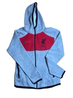 Grå Nike Liverpool hoodie med röd panel - Snygg grå hoodie från Nike med röd panel och Liverpool FC-logga på bröstet. Dragkedja framtill, två fickor och svart Nike-märke. Mjuk och bekväm, perfekt för dig som älskar fotboll och vill ha en sportig stil.