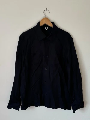 Mörkblå Arket överskjorta - Stilren mörkblå Arket överskjorta med klassisk krage och knäppning framtill. Skjortan har långa ärmar och två bröstfickor. Perfekt att bära öppen över en t-shirt eller stängd för en clean look. 6R5