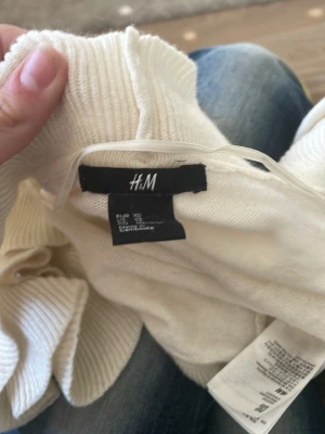 Vit volangkofta från H&M - Söt vit kofta från H&M med breda volangdetaljer längs hela kanten och ärmsluten. Koftan är öppen framtill och har en mjuk, stickad känsla. Perfekt för att lyfta en enkel outfit med lite extra stil.