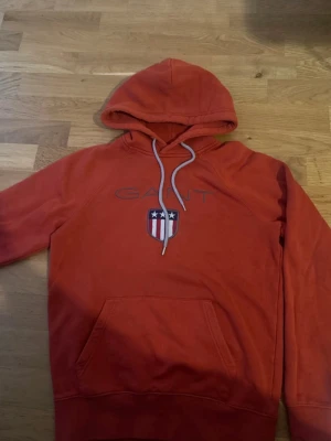 Röd hoodie från Gant med logga - Snygg röd hoodie från Gant med stor broderad logga och sköld på bröstet. Klassisk känguruficka framtill och justerbar huva med vita snören. Perfekt för dig som gillar streetwear och vill ha en bekväm och stilren look.