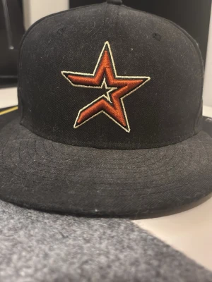 Svart keps med Astros logga - Cool svart keps från New Era med Houston Astros broderad logga i orange och vitt framtill. MLB-logotypen är broderad på baksidan. Klassisk flat brim och strukturerad form, perfekt för dig som gillar sport och streetstyle.