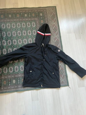 Svart Moncler vindjacka med huva - Snygg svart vindjacka från Moncler med huva och dragkedja. Jackan har vita och röda detaljer på huvan, två fickor med knappar och Moncler-logga på ärmen. Tillverkad i lätt, vindtätt material som passar perfekt för blåsiga dagar.