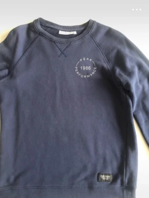 Mörkblå sweatshirt Peak Performance - Mörkblå sweatshirt från Peak Performance med tryck '1986' och logga på bröstet. Klassisk rund halsringning och raglanärmar. Mjuk bomullsmix och diskret etikett nertill. Perfekt för en avslappnad och sportig stil.