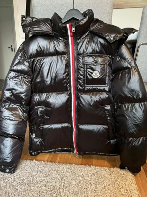 Svart glansig skän rätt tunn kan används t sommaren med  - Snygg svart dunjacka från  med glansigt material, stor huva och rödvit rand längs dragkedjan. Jackan har en bröstficka med Moncler-logga och två sidofickor. Perfekt för kalla vinterdagar och riktigt trendig look.