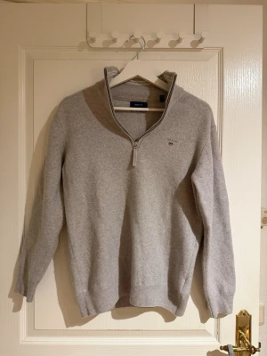 Beige halvzip-tröja från Gant - Beige stickad tröja från Gant med halv dragkedja och liten broderad logga på bröstet. Tröjan har ribbade muddar vid ärmslut och nederkant samt en klassisk, avslappnad passform. Perfekt för lager på lager och enkel att matcha. Storlek L men skulle säga mer som S