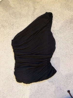 Svart oneshoulder topp från Zara - Säljer en svart oneshoulder topp från Zara. Toppen har en draperad look med snygga veck och är tillverkad i ett lätt, mjukt material. Tröjan är i storlek S och säljer den för den är för liten för mig. Den har tyvärr ett hål på sidan av magen men syns inte så mycket och enkelt att fixa. 