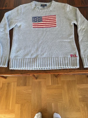 Grå stickad tröja Polo Ralph Lauren - Grå stickad tröja från Polo Ralph Lauren med amerikansk flagga i rött, vitt och blått på bröstet. RL-broderi i rött vid nederkanten. Hör av dig vid funderingar 